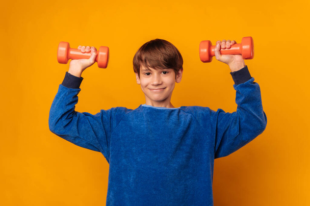 Strong handsome boy is lifting up two orange dumbbells over yellow backdrop in a studio. - Foto, imagen