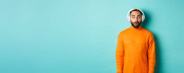 Amazed adult man listening music in headphones, looking at camera impressed with sound, standing over turquoise background. - 写真・画像