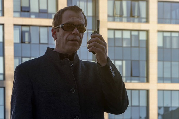 An elderly man in dark glasses speaks on a walkie-talkie against a blurry background of a glass building. - Foto, Bild