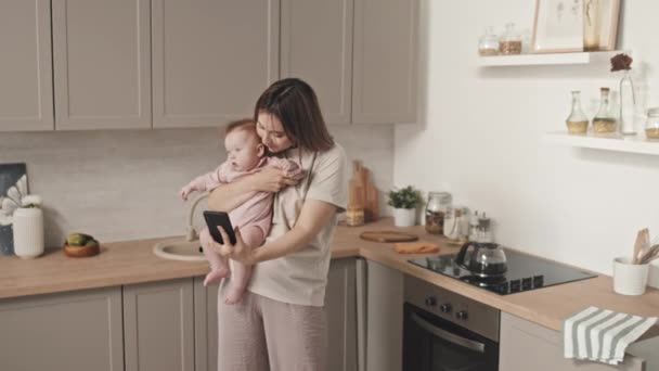 Mittellange braunhaarige junge Frau mit süßer kleiner Tochter, die tagsüber in der Küche steht und Videotelefonie per Smartphone hat - Filmmaterial, Video