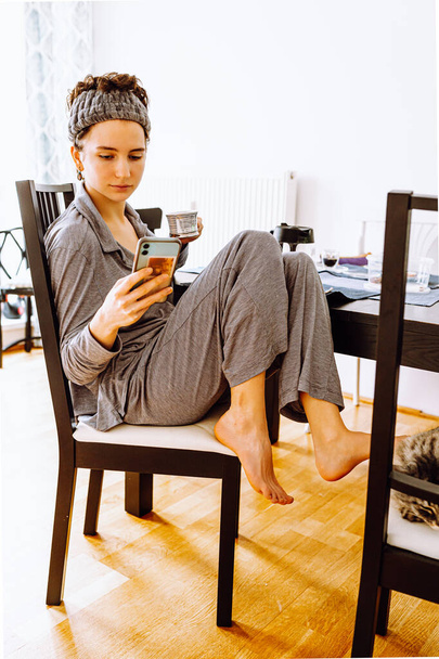teenage girl with bandage on head, in gray pajamas, sits on chair, tucking bare legs under her, drinking coffee, holding smartphone, in sunny morning dining room, domestic cat sits on chair next. Morning positive in pajamas - Fotoğraf, Görsel