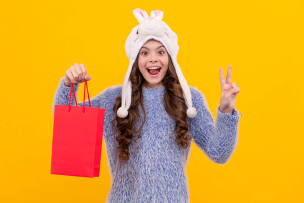 birthday present package. surprised kid with purchase in hat. boxing day. teen girl in knitwear on yellow background. child buy gift in shopping bag. black friday discount. christmas sale. - Foto, Bild