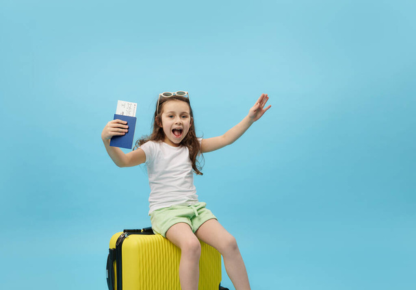 Cheerful little traveler passenger tourist, child girl holding a boarding pass, sitting on yellow suitcase, expecting the upcoming flight, isolated blue background. Trip Travel Summer holidays concept - Foto, Imagem