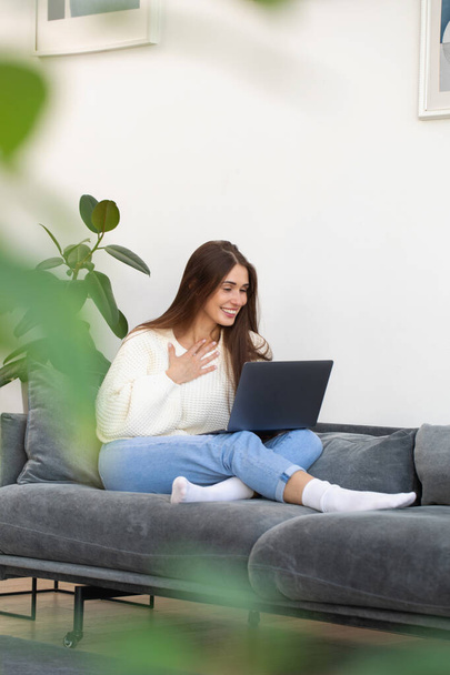 A young woman looks at a laptop monitor and smiles, sitting on the sofa in her living room surrounded by house plants. - Foto, imagen