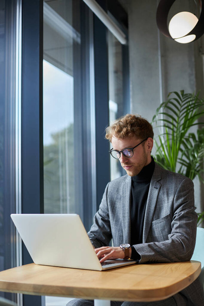 Caucasian confident young man in formal suit and eyeglasses, entrepreneur, sales manager typing on laptop keyboard while telecommuting from a modern office. People, business and freelance work concept - Fotó, kép