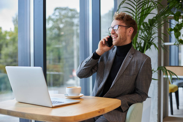 Handsome Caucasian young confident male entrepreneur sitting at desk with laptop, using mobile phone, discussing business project with partner or investors while telecommuting from an office interior - Фото, изображение