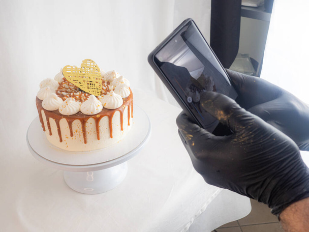 chef pastry baker making a reel of a salty caramel frosted dripping cakes with meringues and chocolate heart topper with smarphone photo - Valokuva, kuva