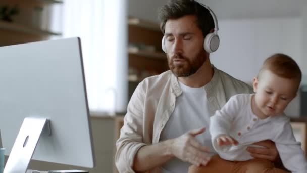 Remote work and parenthood. Young busy father businessman video chatting with partners via pc and carrying his active newborn baby on hands, working from home, tracking shot, free space - Filmati, video