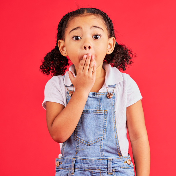 Shocked kid, portrait and hand on mouth in secret, oops and mistake facial expression on isolated red background. Child, little girl and surprised face in gossip, news or emoji at studio announcement. - Fotó, kép