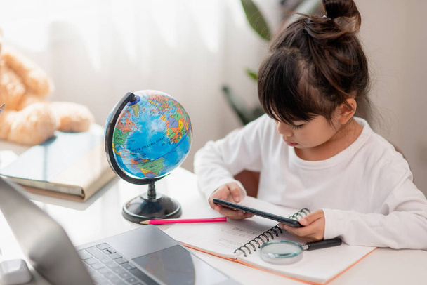 Asian little girl  using a phone close up, distracted from studying, sitting at a table with notebooks, a pretty child having fun with a smartphone, watching the webinar, homeschooling - Photo, Image