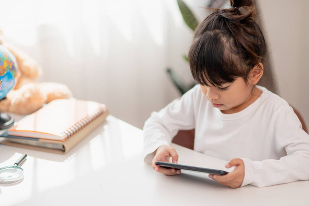 Aasialainen pikkutyttö käyttää puhelinta lähikuva, hajamielinen opiskelusta, istuu pöydässä muistikirjojen kanssa, kaunis lapsi pitää hauskaa älypuhelimen kanssa, katselee webinaaria, kotiopetusta - Valokuva, kuva