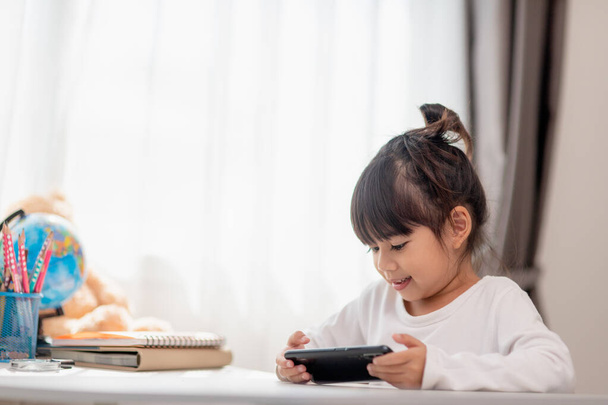 Asiatisches kleines Mädchen mit Handy aus nächster Nähe, abgelenkt vom Lernen, an einem Tisch mit Notizbüchern sitzend, ein hübsches Kind, das Spaß mit dem Smartphone hat, das Webinar anschaut, Hausaufgabenbetreuung - Foto, Bild