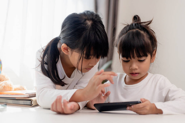 Çocukları ve aletleri kavrayın. İki küçük kız kardeş telefona bakar ve gülümser. Akıllı telefon videoları var, öğreniyorlar, oyunlar oynuyorlar, internette konuşuyorlar. Çocuklar için internet. Ebeveyn kontrolü - Fotoğraf, Görsel