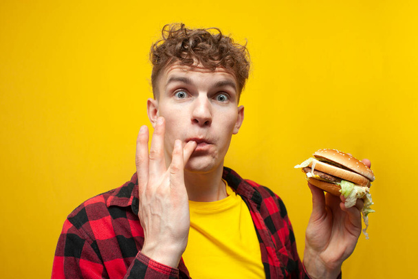 young surprised guy student eats a big tasty burger on a yellow background and licks the sauce on his finger, a man eats fast food and looks at the camera - Photo, image