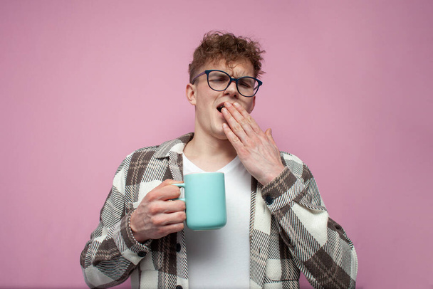 втомлений сонний молодий хлопець в окулярах тримає чашку кави і повії на рожевому фоні, невтомний студент з напоєм покриває рот рукою
 - Фото, зображення