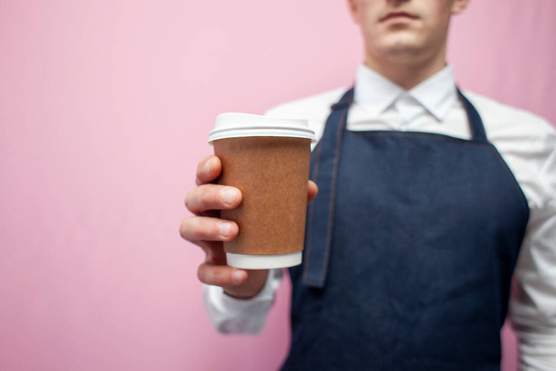 白いシャツとエプロンの若いバリスタがピンクの背景にコーヒーカップを持ちバリスタの労働者の手による紙コップのクローズアップ - 写真・画像