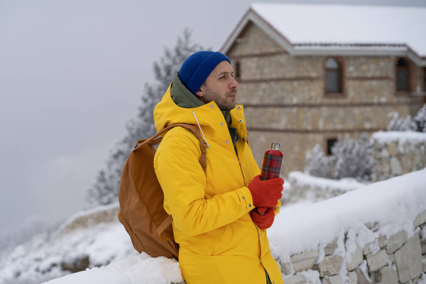 Thoughtful man walks with hot drink in thermal bottle thinks question, dreaming of future at winter frosty day. Relaxed male stands snow outdoor in yellow clothes for cold weather looks into distance - Photo, Image