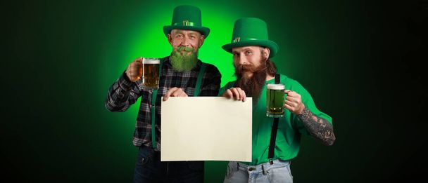 Bearded men with blank poster and glasses of beer on dark green background. St. Patrick's Day celebration - Fotografie, Obrázek