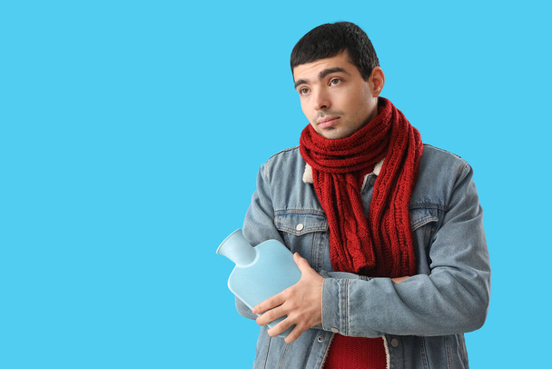 Young man warming his arm with hot water bottle on blue background - Valokuva, kuva