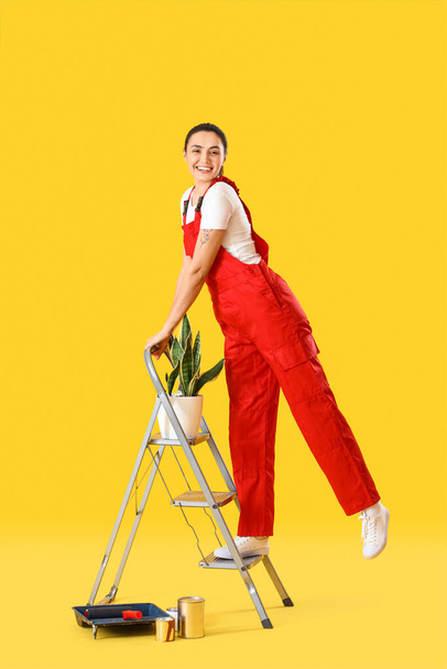Young woman with houseplant, ladder and paint cans on yellow background - Фото, зображення