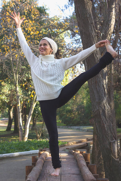 Smiling middle aged woman in yoga standing balance Parsva Utthita Hasta Padangusthasana. Female mature yogi on wooden path with extended hand to big toe pose. Fall season, sport activity in the park - Foto, afbeelding