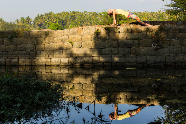 Yogi woman in purvottanasana pose on top of rocks wall with reflection on river by Srirangapatna, Karnataka, India. Nature connection concept - 写真・画像