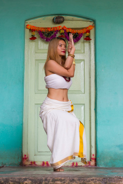 Indian yogi girl in traditional Kerala clothes in garudasana pose. Young woman practices eagle yoga pose, with a white door on turquoise blue background in Mysore, India - Fotografie, Obrázek