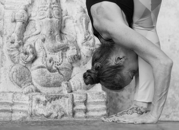 Woman practicing yoga by Ganesha sculpture with head between her legs and hands on the floor, in uttanasana pose in Srirangapatna temple, Karnataka, India. Black and white photography - Fotografie, Obrázek