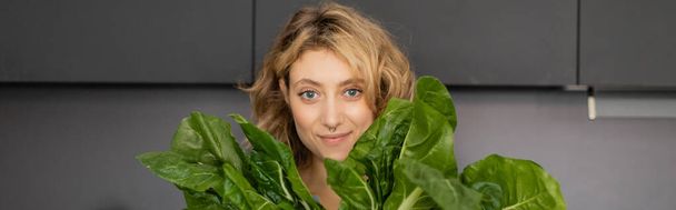 przekłuta młoda kobieta uśmiechnięta i trzymająca liście zielonej kapusty w kuchni, sztandar  - Zdjęcie, obraz