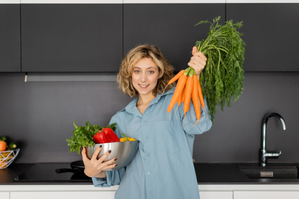 明るい若い女性が野菜と新鮮なニンジンをキッチンで持っています  - 写真・画像
