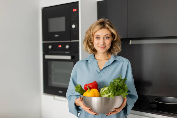 pleased young woman holding bowl with fresh vegetables in kitchen  - Fotografie, Obrázek