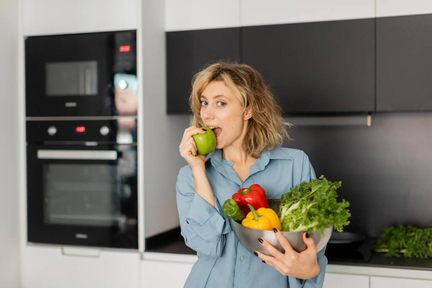 blonde woman biting apple and holding bowl with fresh vegetables in kitchen  - 写真・画像