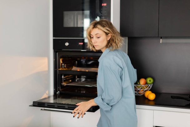 giovane donna dai capelli ondulati che utilizza un forno moderno mentre cucina in cucina  - Foto, immagini