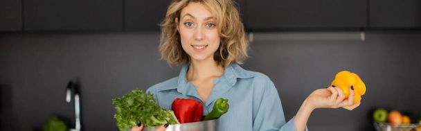 joyeuse jeune femme aux cheveux ondulés tenant bol avec des légumes biologiques dans la cuisine, bannière  - Photo, image