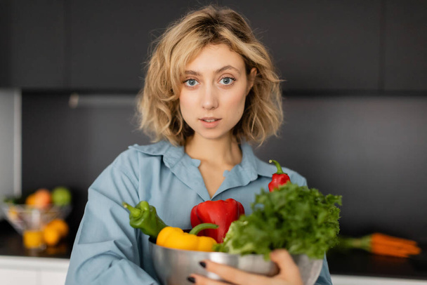 blonde young woman with wavy hair holding bowl with organic vegetables in kitchen  - Foto, Imagen