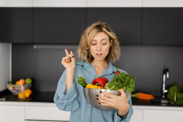 блондинка молода жінка з хвилястим волоссям тримає миску з органічними овочами і показує знак ідеї
  - Фото, зображення