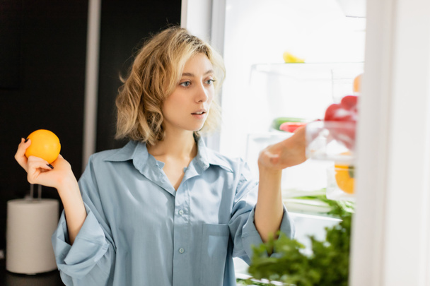 thoughtful young woman holding orange and looking at refrigerator in kitchen  - Foto, Imagen