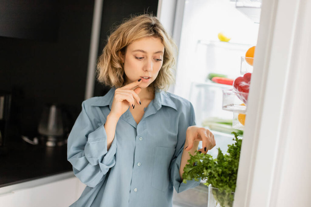 pensive jonge vrouw op zoek naar groen in open koelkast  - Foto, afbeelding