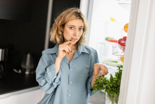 задумчивая молодая женщина смотрит в камеру рядом с открытым холодильником на кухне  - Фото, изображение