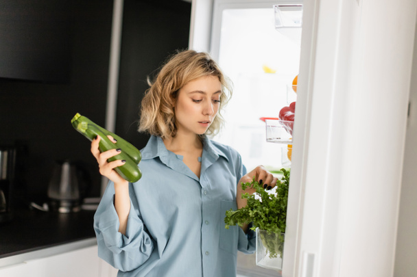mujer joven rubia en camisa azul mirando a la vegetación mientras sostiene calabacín cerca de nevera abierta en la cocina  - Foto, imagen