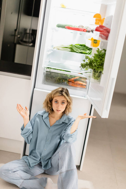 σύγχυση νεαρή γυναίκα κάθεται κοντά άνοιξε ψυγείο με φρέσκα προϊόντα στην κουζίνα  - Φωτογραφία, εικόνα