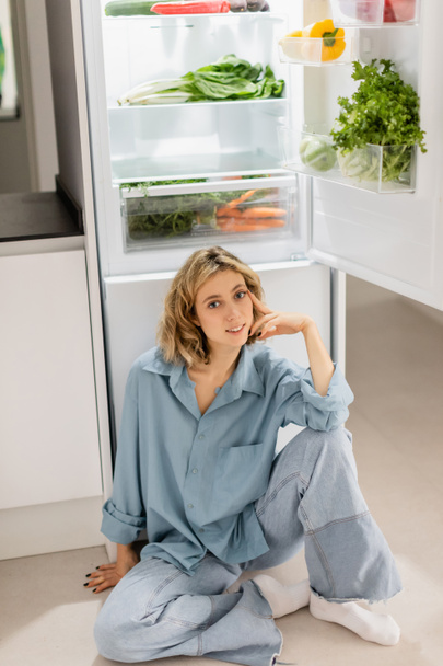 счастливая молодая женщина сидит рядом с открытым холодильником со свежими продуктами на кухне  - Фото, изображение