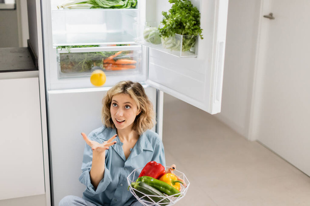 young woman sitting near opened refrigerator with fresh vegetables while throwing lemon in air  - Foto, imagen
