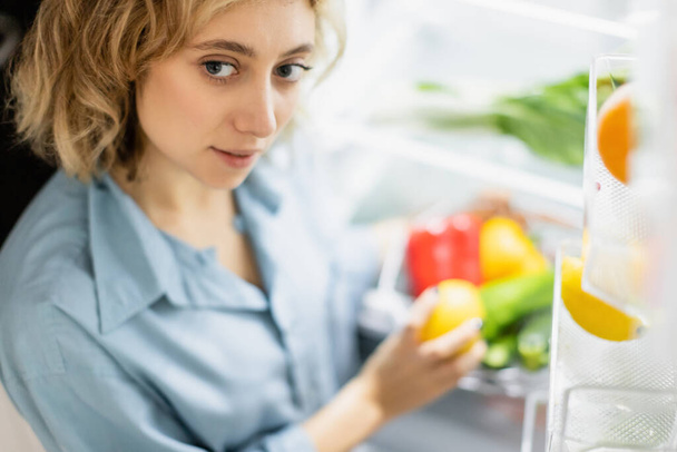 Blick aus der Vogelperspektive auf eine junge Frau, die in der Nähe eines geöffneten Kühlschranks steht  - Foto, Bild