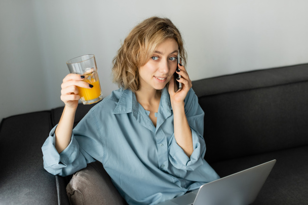 alegre freelancer con cabello ondulado sosteniendo vaso de jugo de naranja mientras habla en el teléfono inteligente cerca de la computadora portátil  - Foto, imagen