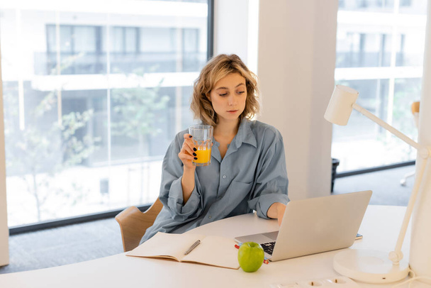 young woman with wavy hair holding glass of orange juice near laptop on desk  - Foto, imagen