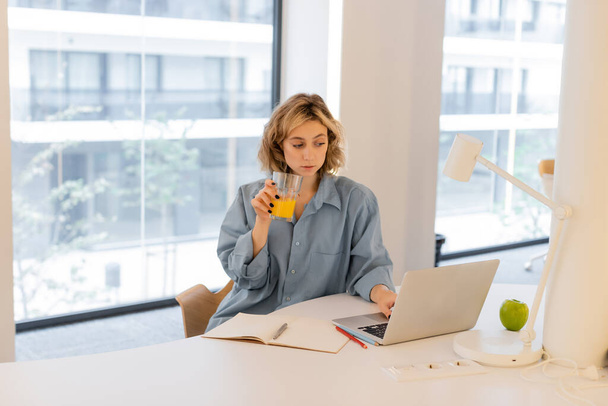 mujer joven con cabello ondulado sosteniendo un vaso de jugo de naranja fresco mientras usa la computadora portátil  - Foto, imagen