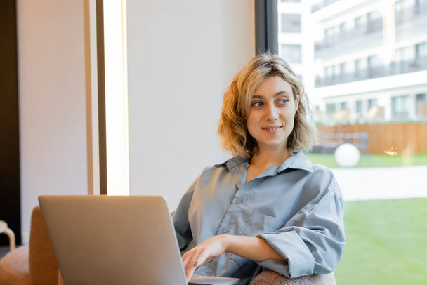 happy blonde woman with wavy hair looking at window and sitting with laptop  - Photo, Image