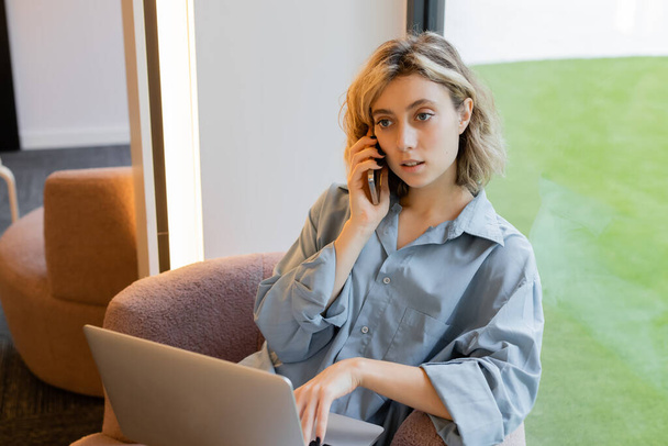 young blonde woman with wavy hair talking on smartphone while using laptop in cafe  - Foto, immagini