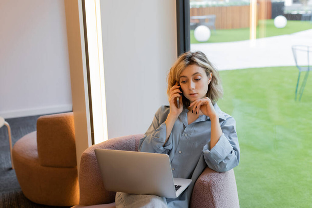 blonde freelancer with wavy hair talking on smartphone while using laptop in cafe  - Valokuva, kuva